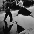 Un uomo dà una donna una mano mentre vola  su una grande pozzanghera sul marciapiede, 1960