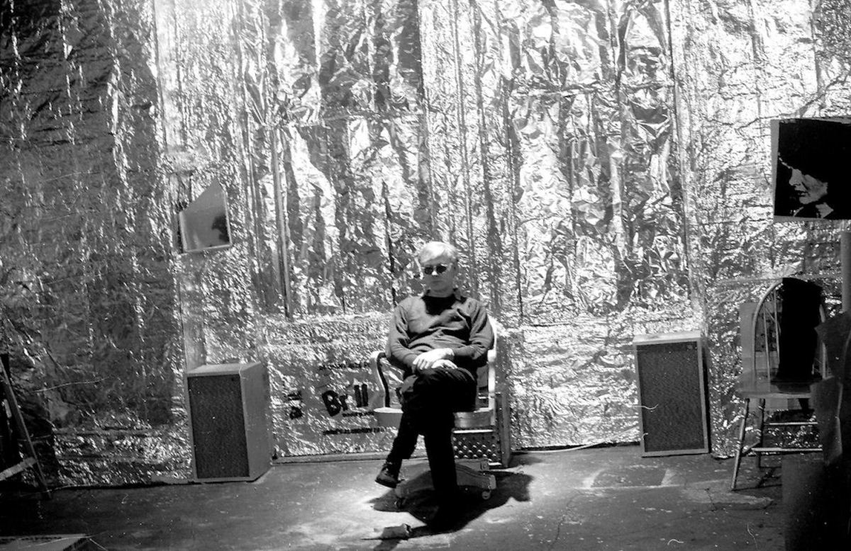 Andy Warhol seduto su una sedia alla Factory con la tela di Jacqueline Kennedy, le scatole Brillo e la parete d'argento. New York NY US 1964 Billy Name / NOvoWorks